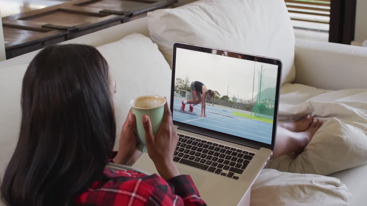 Composite of woman sitting at home holding coffee watching athletics running event on laptop