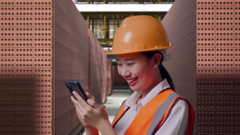 close up side view of asian female engineer with safety helmet using smartphone while standing with red brick packed in stacks are stored