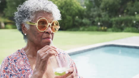 una mujer afroamericana de edad avanzada feliz con gafas de sol junto a la piscina bebiendo cócteles, copiando espacio, cámara lenta