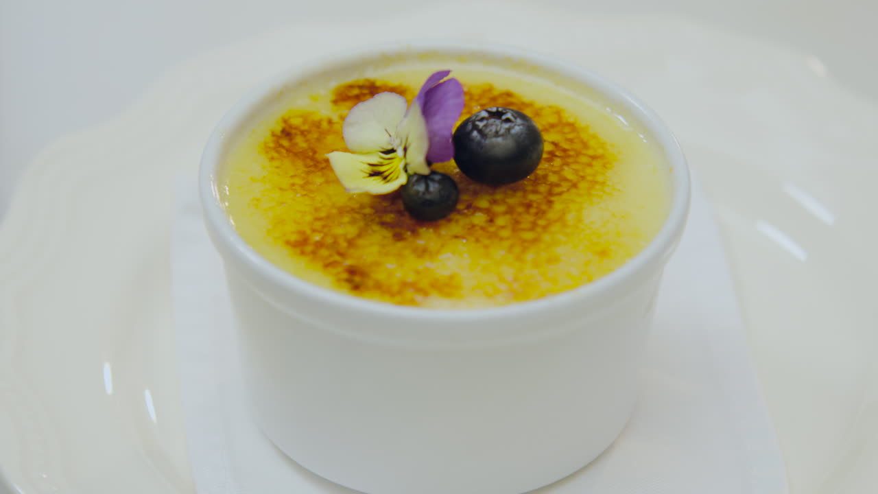 Close-up of a completely golden creme brulee, decorated with an edible flower and fresh blueberries, while a spoon cuts its caramelized surface