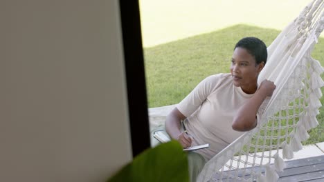 feliz mujer afroamericana sentada en una hamaca en la terraza, escribiendo en un cuaderno, cámara lenta