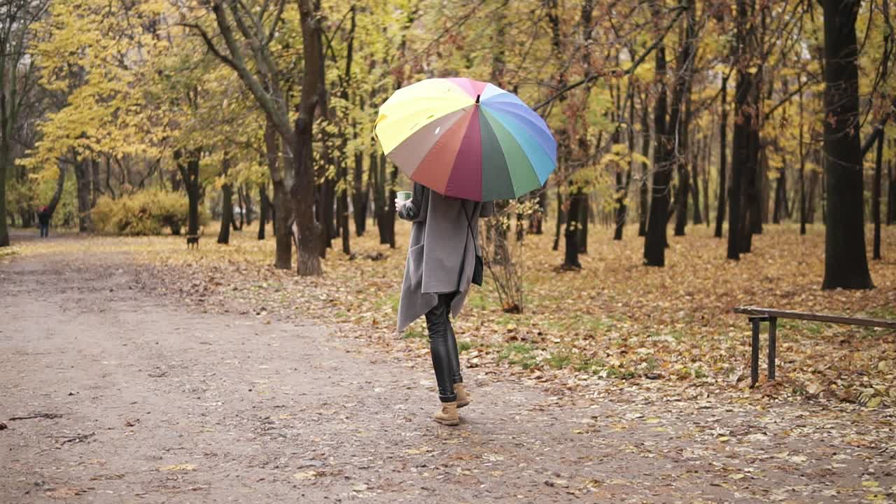 vista trasera de una mujer joven de cabello rojo caminando en el parque de otoño con un colorido paraguas arco iris y sosteniendo una taza de papel con café. niña con abrigo cálido disfrutando del clima de otoño fresco con una copa de bebida caliente