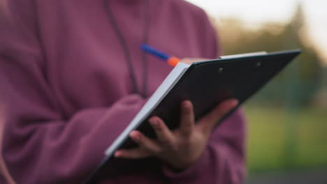 primer plano de una persona con una camiseta marrón escribiendo notas al aire libre, sosteniendo un portapapeles con una pluma, con el fondo borroso del campo deportivo y la cancha