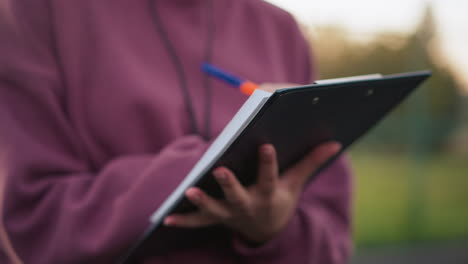 close-up of person in maroon top writing notes outdoors, holding clipboard with pen, with blurred background of sports field and court