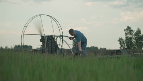 young man in blue shirt leans over paramotor on grassy field at dusk starting engine causing fan blades to spin rapidly within metal cage against open farm sky and ready for flight