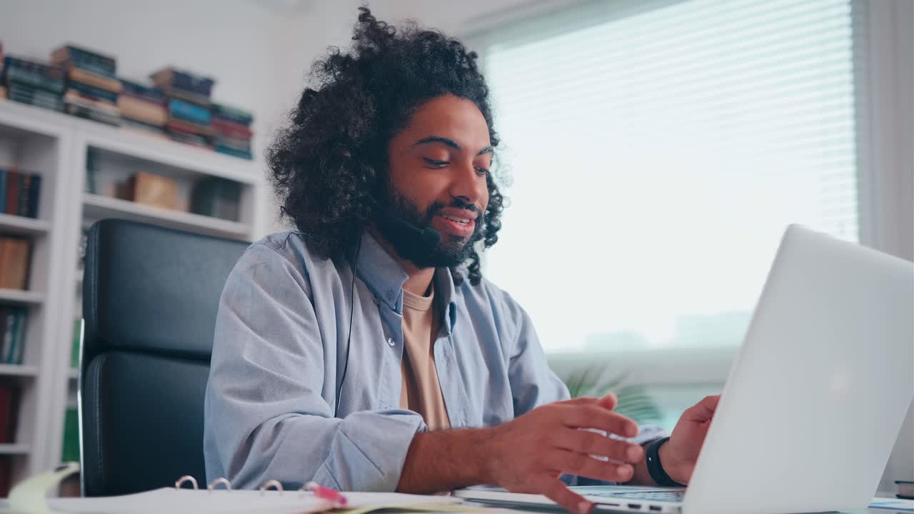 Enthusiastic young arabian man smiling talking over headset looking in laptop
