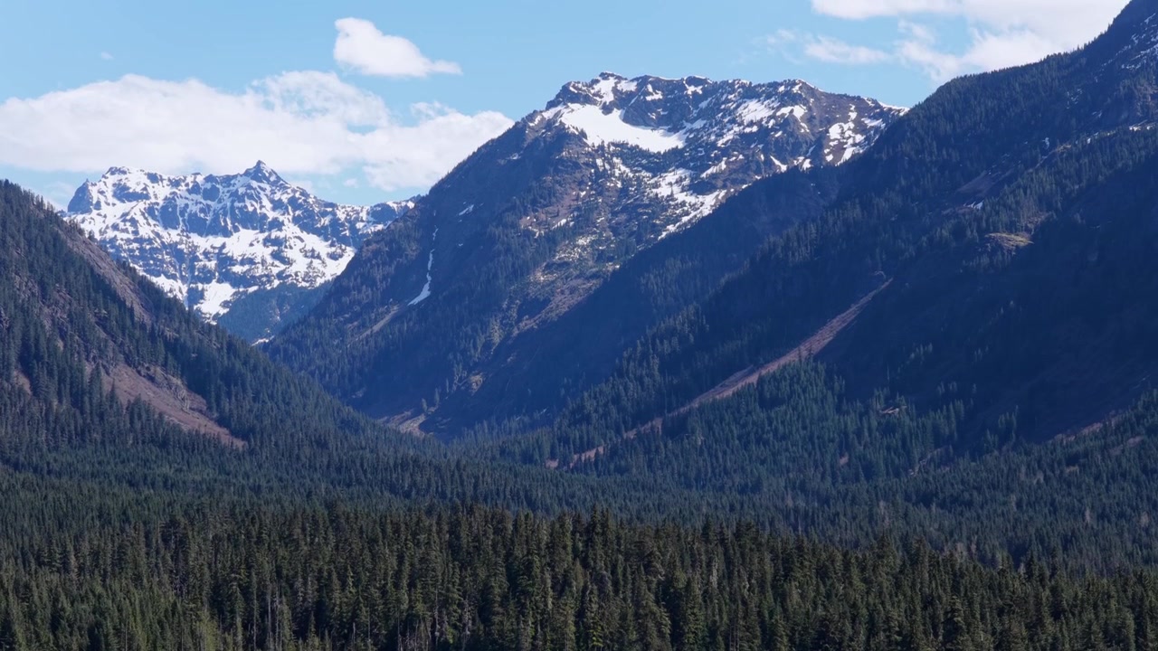 Passo Snoqualmie con lago alpino incastonato in una fitta valle di foresta sempreverde, cime innevate e pendii rocciosi che si ergono ripidamente sopra un prato umido sotto un cielo blu. Washington, USA. Vista aerea