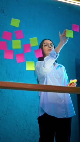 lady in white shirt and black trousers puts stickers on the glass wall. placing memo notes on the board in office. vertical video.