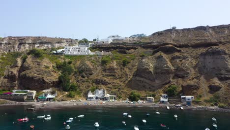 Vista-Estática-Aérea-De-La-Isla-Griega,-Acantilado-Rocoso-Con-Villas-Y-Barcos-De-Pesca-En-El-Mar-Mediterráneo-En-La-Playa-De-Santorini,-Grecia