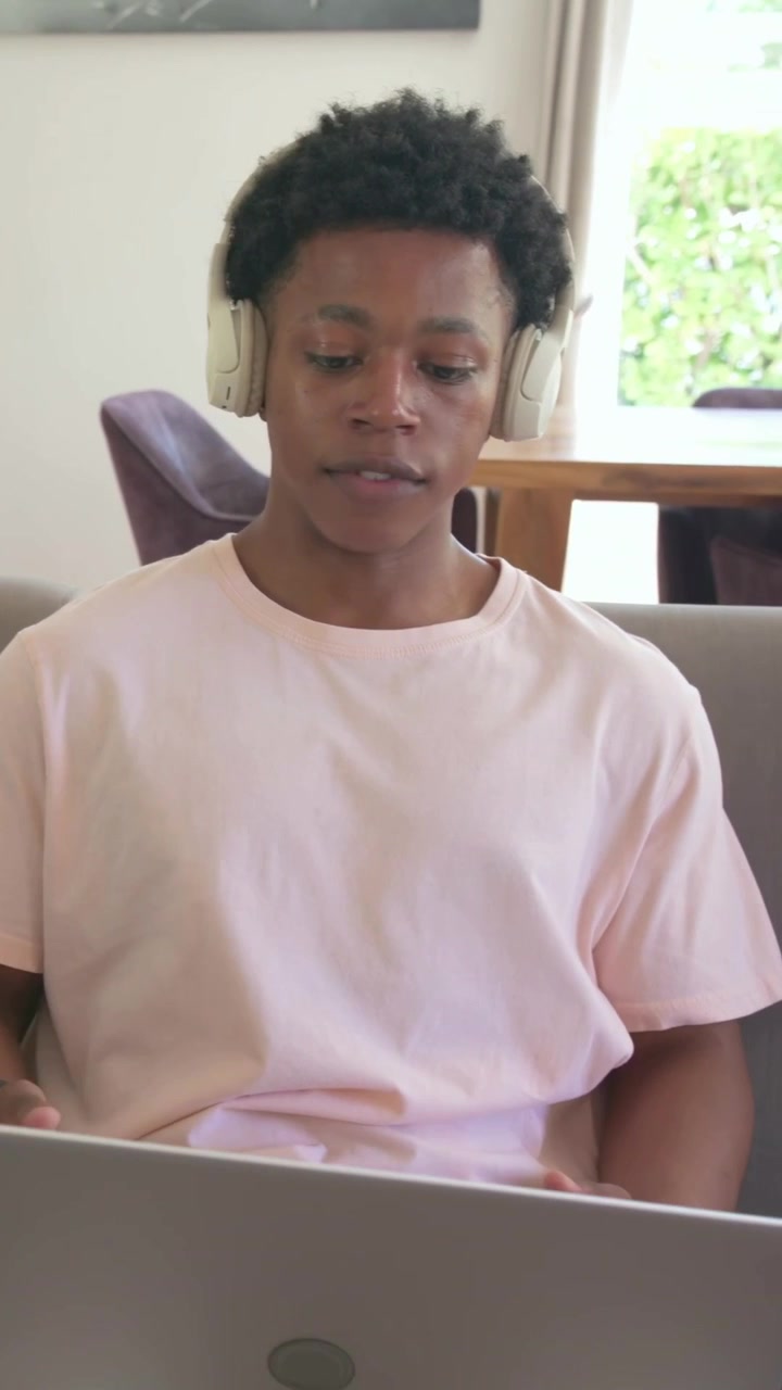 Vertical video: Joining meeting, man wearing pink tee and headphones gesturing on couch with laptop