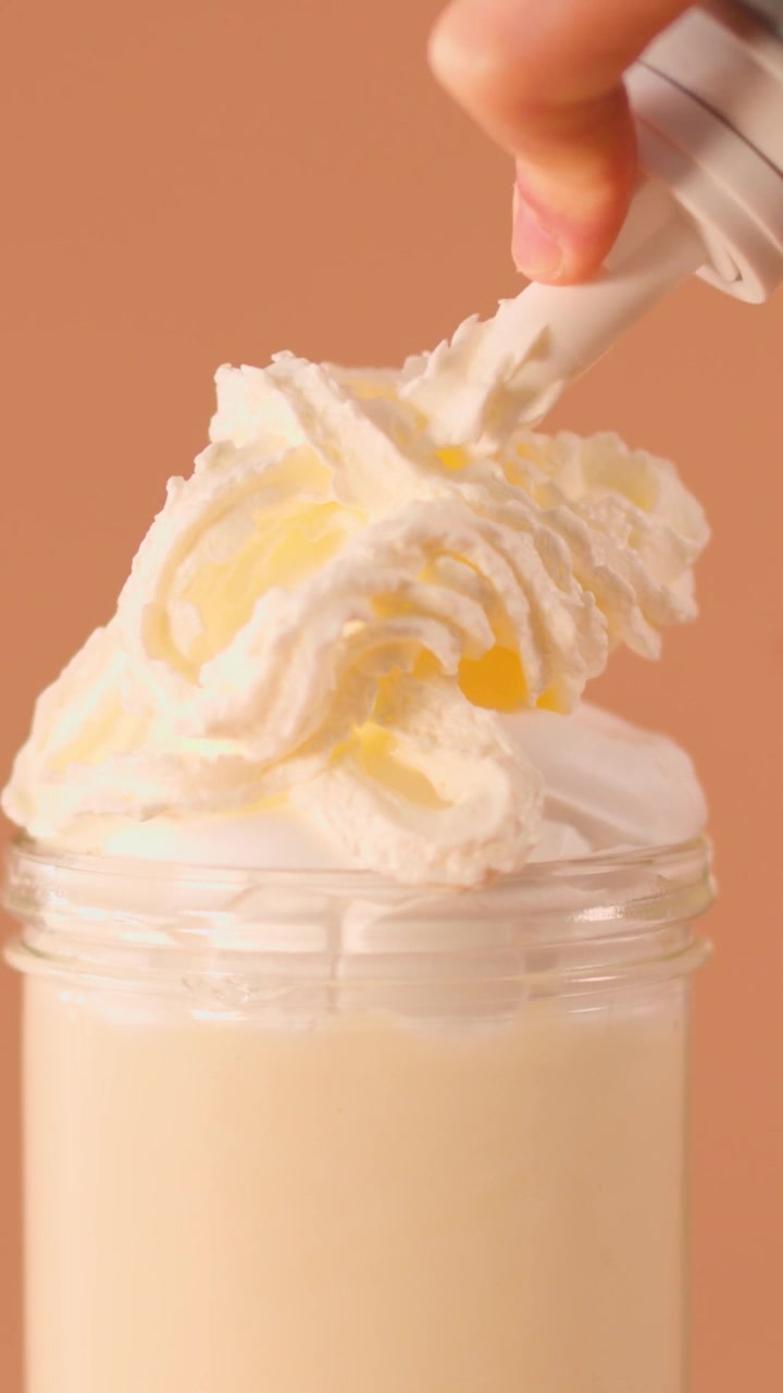 Primo piano di una mano che guarnisce un cremoso milkshake alla vaniglia con spesse spirali di panna montata.