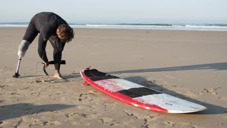 tiro largo de un surfista masculino con una pierna ortopédica poniéndose la correa de la tabla de surf