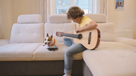 blond boy with curly hair playing the guitar sitting on the couch, next to him is his dog lying