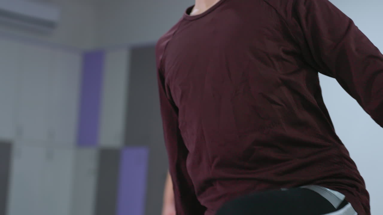 niño joven con gafas estirando el brazo mientras se equilibra en una pierna y trayendo la pierna hacia adelante en una postura enfocada, con vista parcial de otro niño realizando un movimiento similar en un gimnasio brillante