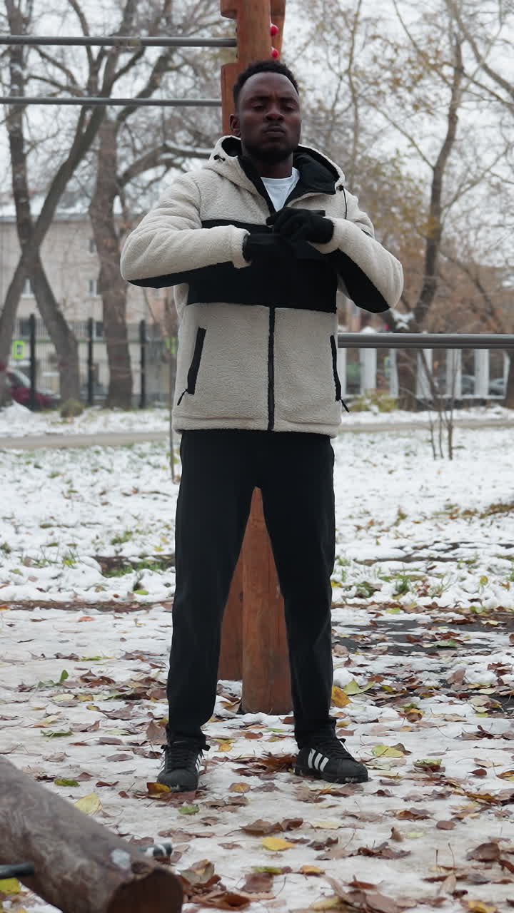 niño al aire libre realizando estiramientos de brazos en invierno. la nieve cubre el suelo, rodeado de equipos de entrenamiento y árboles en el fondo
