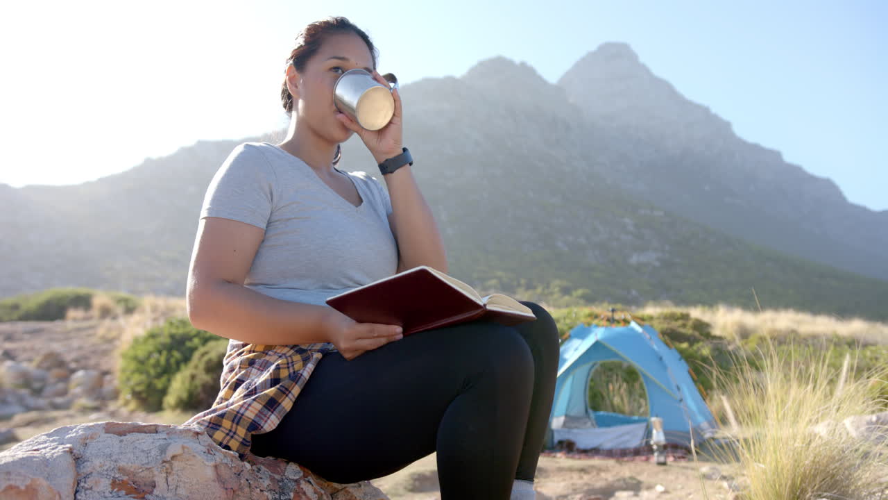 Donna che beve da una tazza e legge un libro mentre è seduta vicino alla tenda
