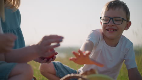 a cheerful child wearing glasses and a white shirt reaches out to grab something from a basket while his mom, holding a red apple, playfully taps his hand to stop him