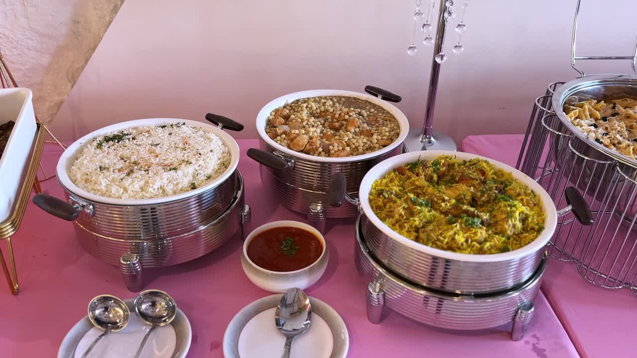 Buffet catering interno con diverse pietanze calde tra cui riso bianco, riso speziato con carne, stufato di ceci e pasta cremosa serviti in scaldavivande in acciaio inossidabile su un tavolo decorato