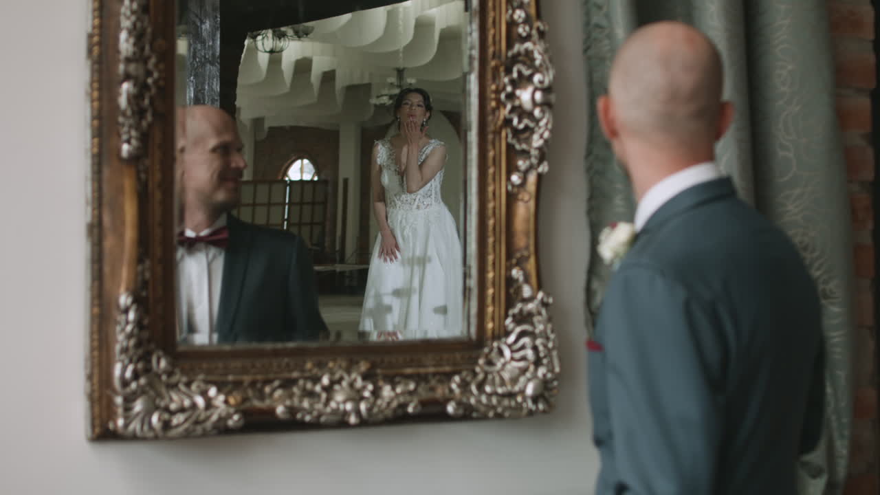 An elegant groom in a suit and bow tie tenderly gazes at his bride's reflection in the mirror as she shows off her wedding dress. He lovingly blows her a kiss.