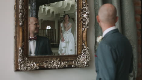 an elegant groom in a suit and bow tie tenderly gazes at his bride's reflection in the mirror as she shows off her wedding dress. he lovingly blows her a kiss.