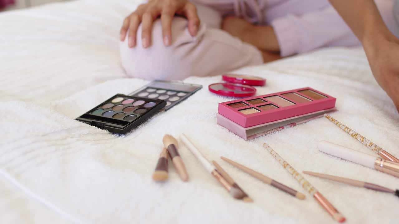 Hands reaching forward are picking up and arranging palettes and brushes on bed preparing makeup