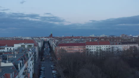 Establisher-shot-of-rooftop-of-traditional-brick-houses-across-street-with-vehicles-parked-and-moving-across-lane-surrounded-with-trees-on-a-cloudy-early-morning-in-Berlin,-Germany