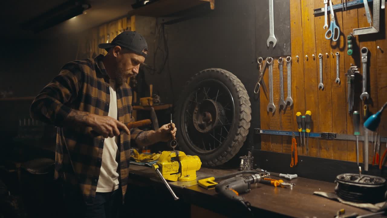 A blond mechanic with a beard in a cap uses a clamp and a drill while working on a workbench in a studio workshop