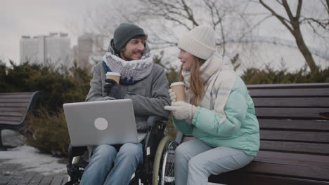 disabled man in wheelchair and his friend watching something funny on laptop computer while drinking takeaway coffee at urban park in winter