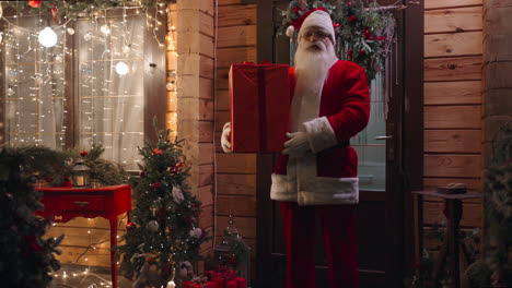 cámara de zoom santa claus se encuentra fuera de la casa al aire libre en invierno con un regalo y mira a la cámara