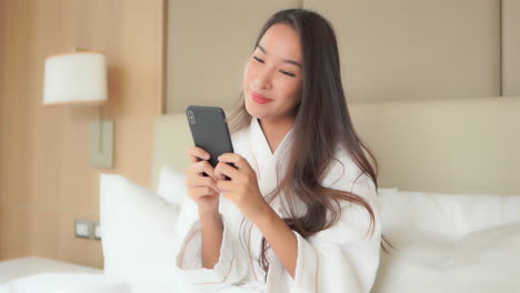 a medium close of a pretty young woman sitting on the bed in her luxury hotel suite while wearing a resort bathrobe, enjoys communicating through her smartphone