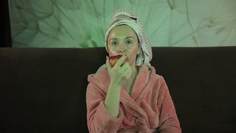 mujer viendo una película nocturna en la televisión, comiendo una manzana. túnica de baño, máscara facial
