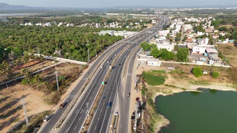 aerial view: multi-lane mysuru highway, busy with traffic, runs alongside palm groves and a large lake. residential areas and development blend with natural elements under a clear sky