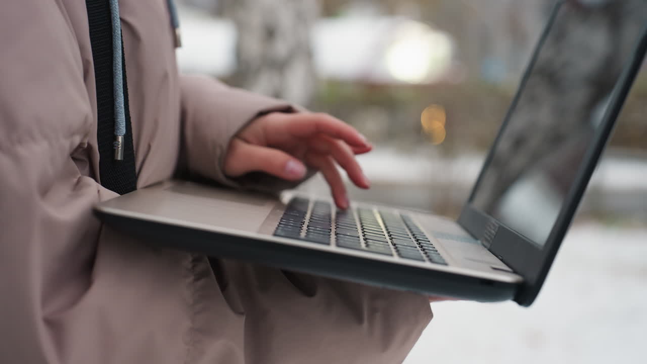 Close-up view of light-skinned female hand operating black laptop while holding it in winter environment, wearing jacket, blurred screen visible, soft background lights and subtle outdoor setting