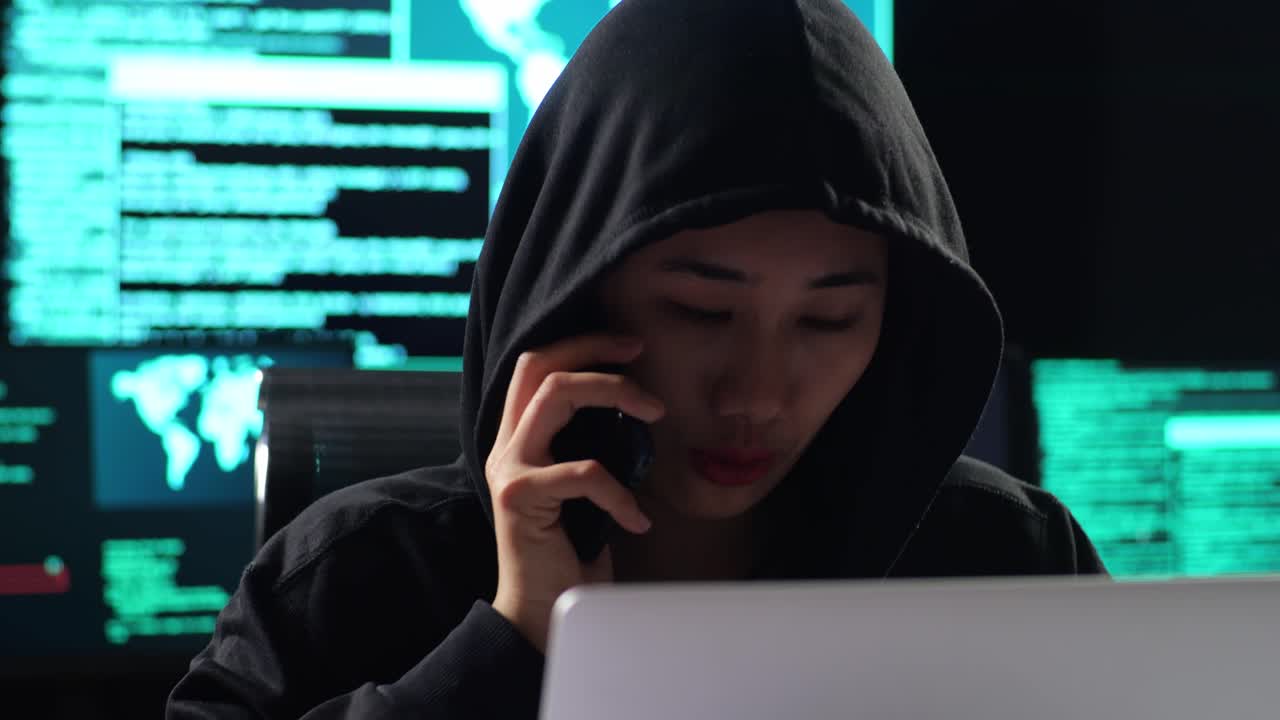Close Up Of Asian Female Hacker In A Hood Talking On Smartphone While Works On A Laptop With Maps And Data On Display Screens Of Multiple Monitors In A Dark Office Room