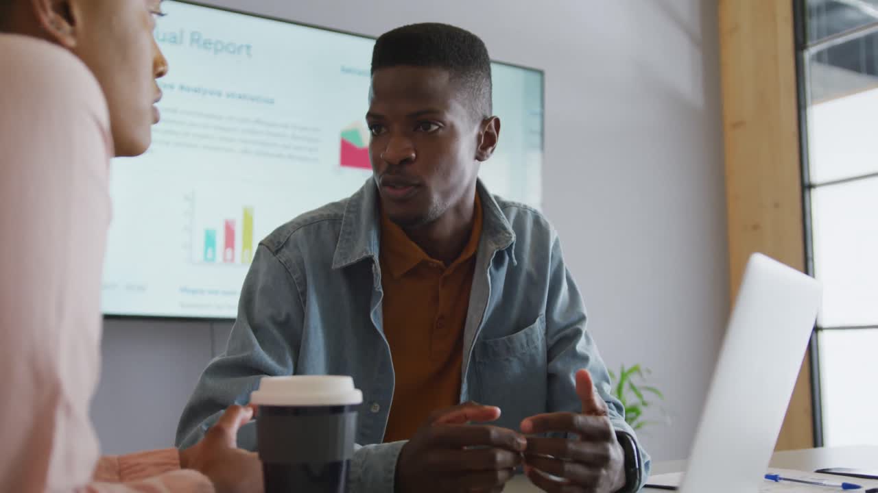 Smiling african american male and female colleague in discussion at meeting using laptop and screen