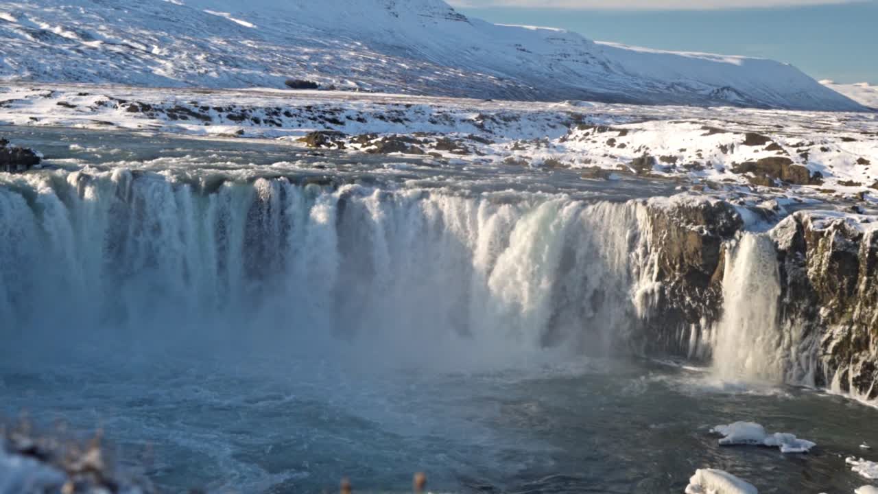 A powerful torrent of water plunges over Goðafoss, Iceland's "Waterfall of the Gods," set against a stunning winter landscape, framed by snow-covered rocks and icy formations