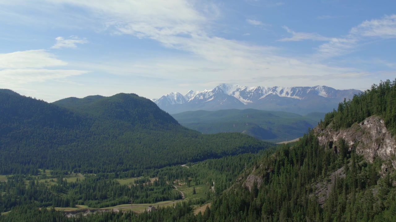 Wide aerial view of green taiga hills and a distant snowcapped mountain range under blue sky near Geyser Lake, Altai Republic, Russia
