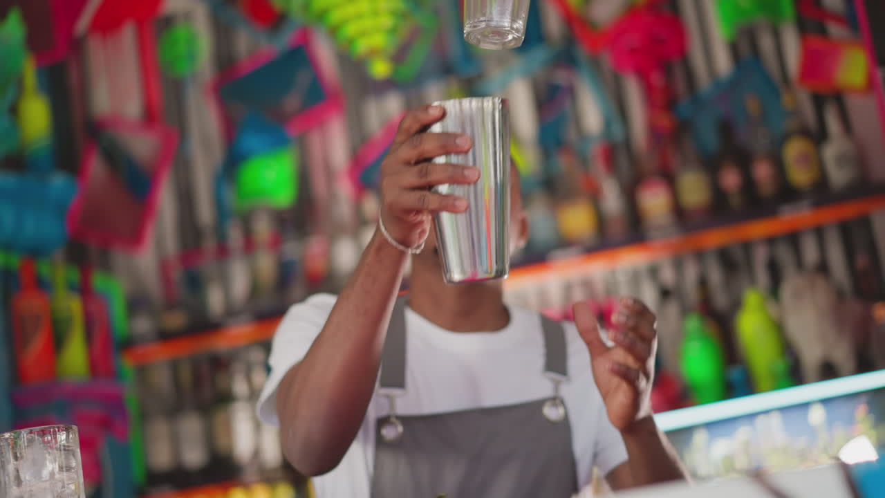 Barman takes shaker and glass in club. Concentrated bartender does tricks with cocktail equipment in bar. African American man works at counter