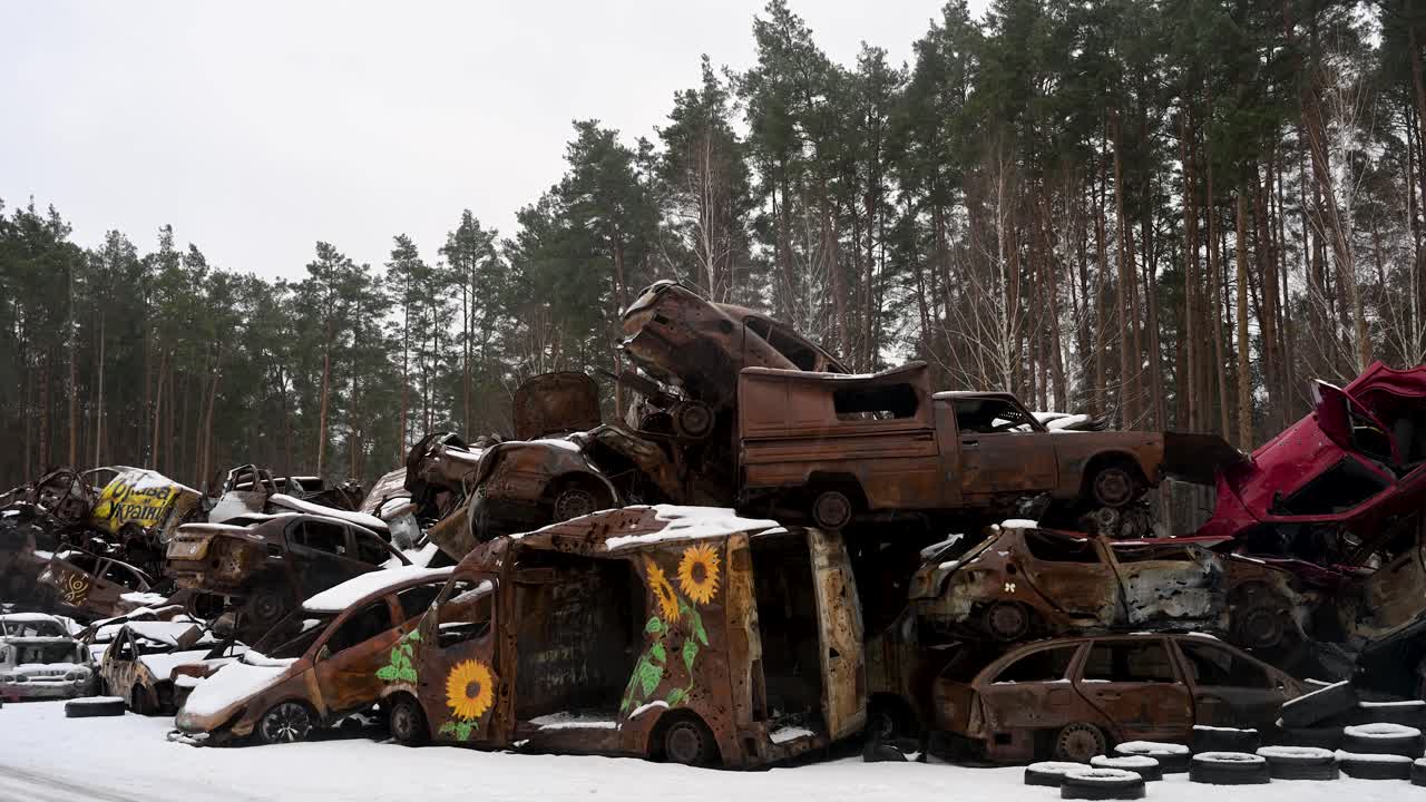On a winter day in Irpin, Ukraine, a pile of burnt and destroyed cars is stacked, with one vehicle featuring sunflower artwork, following intense battles between Ukrainian and Russian forces.