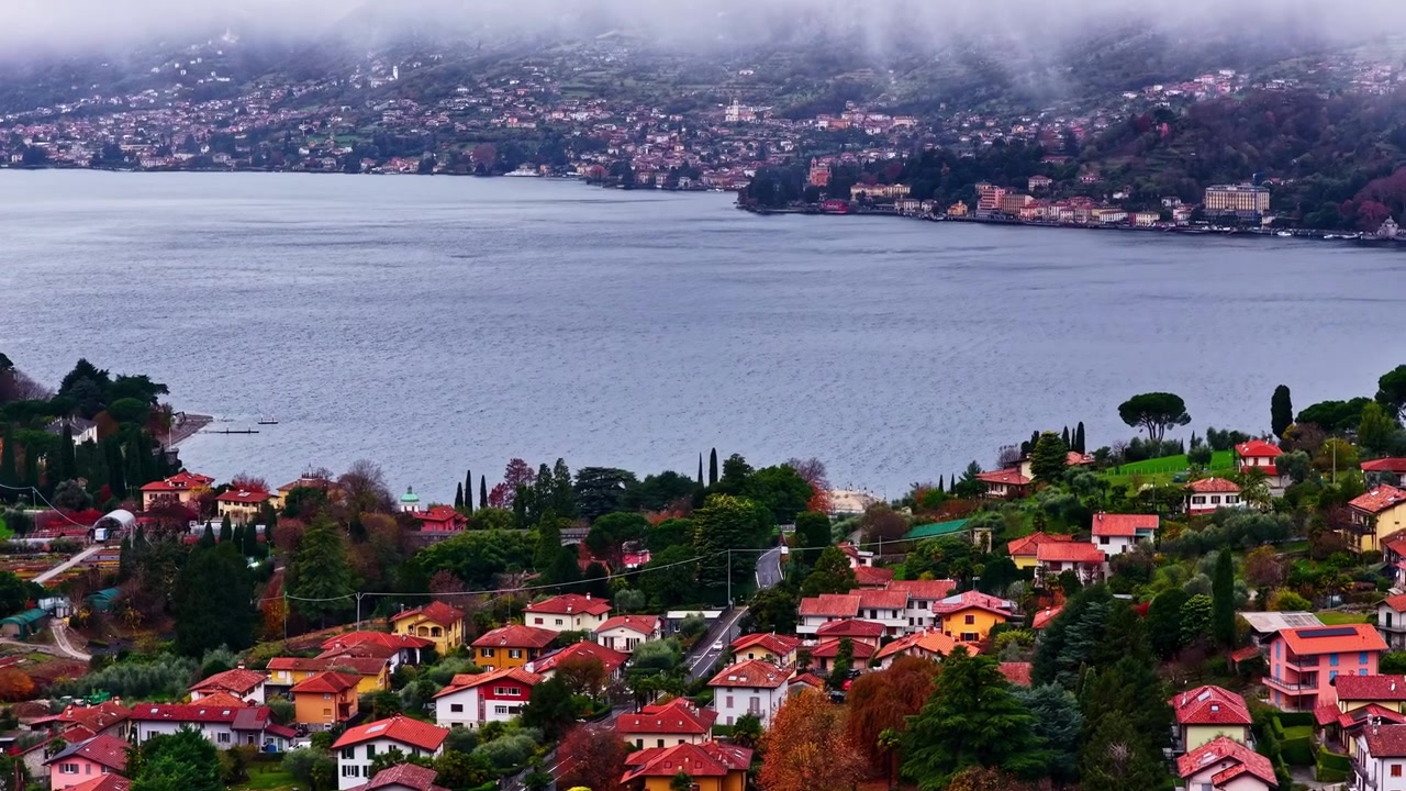 Lago di Como circondato da villaggi con tetti rossi e nebbia bassa sulle montagne vicino a Bellagio, ripresa aerea con drone, carrello a destra e inclinazione verso l'alto.