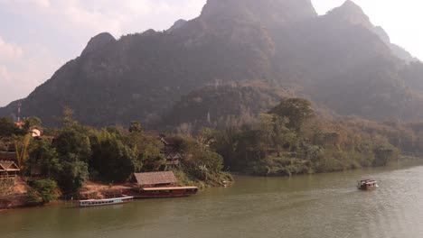 Barco-Flotando-Río-Abajo-Bajo-El-Sol-De-La-Tarde-En-La-Ciudad-Montañosa-De-Nong-Khiaw-En-Laos,-Sudeste-De-Asia