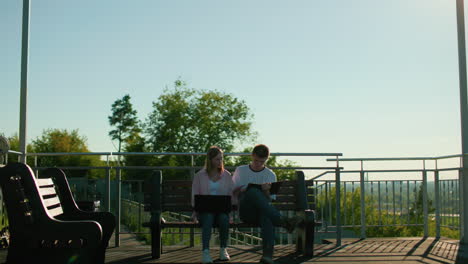 college students studying outdoors with laptop on girl's lap while boy takes notes, she checks his work, surrounded by greenery, light poles, and wooden benches under a serene, sunny environment