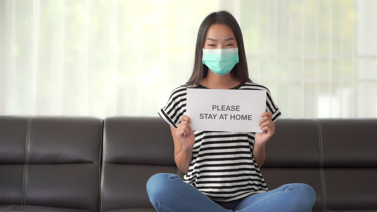 Una bellissima donna asiatica seduta sul divano con una mascherina chirurgica che le copre il viso e che tiene in mano un cartello che incoraggia le persone a restare a casa a causa della pandemia di Covid-19