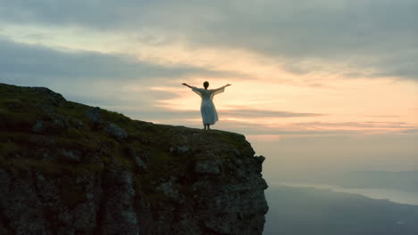person with outstretched arms standing on a cliff edge at sunset