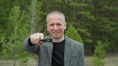 a close-up view of a man in a gray jacket and black shirt standing in front of a green forest background, pointing a pistol, his expression is serious and focused