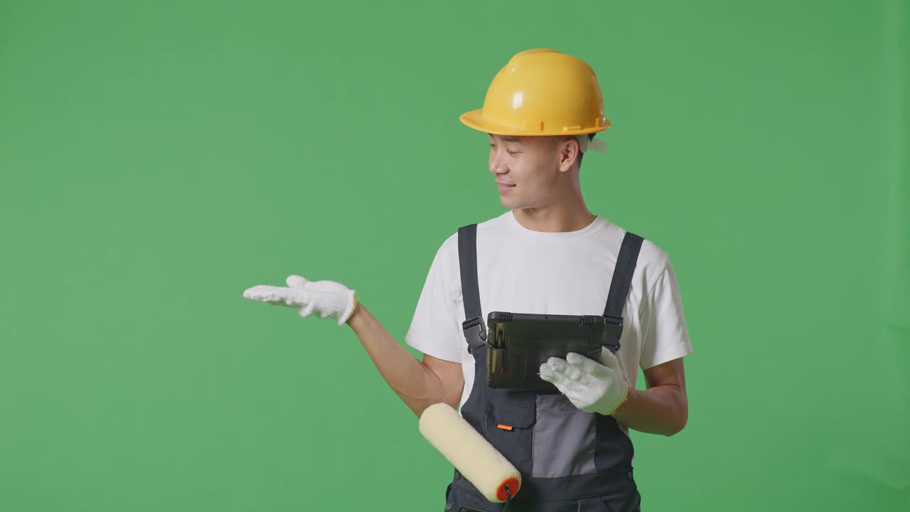 Asian Man Painter Wearing Safety Helmet Using A Tablet And Pointing To Side While Standing In The Green Screen Background Studio
