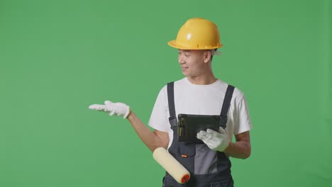 asian man painter wearing safety helmet using a tablet and pointing to side while standing in the green screen background studio
