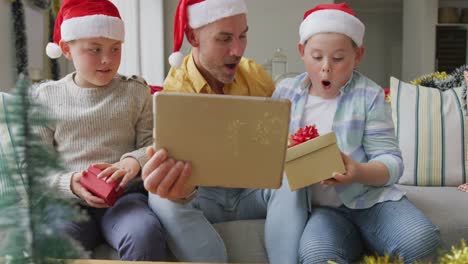 caucasian father and two sons opening gifts having a videocall on digital tablet during christmas