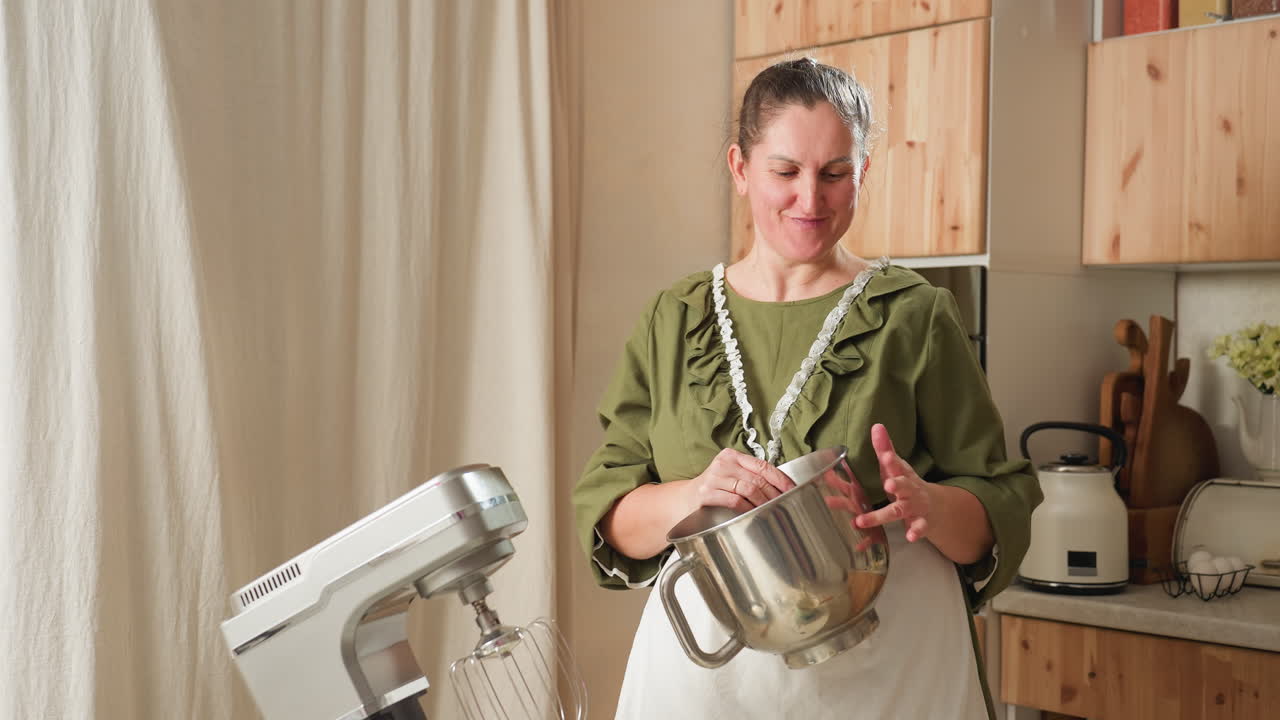 Smiling older woman in green dress and apron cheerfully beats mixing bowl while dancing playfully in cozy kitchen with mixer and cooking utensils in background