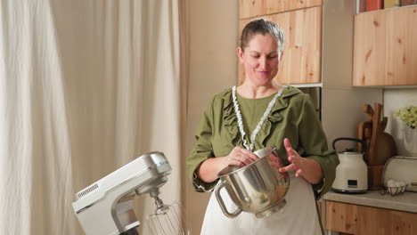 smiling older woman in green dress and apron cheerfully beats mixing bowl while dancing playfully in cozy kitchen with mixer and cooking utensils in background